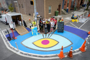 Oliver Allover Eyes community paint day drone birds eye view of participants standing behind eyeball street mural