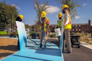 Cecil Community Park: Graham Projects team member texture spraying turquoise pavement art on an asphalt walking track with two people on either side of them shielding the overspray.