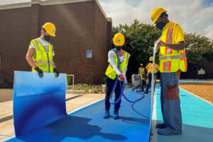Cecil Community Park: Graham Projects team member texture spraying blue pavement art on an asphalt walking track, with two people on either side of her shielding the overspray.