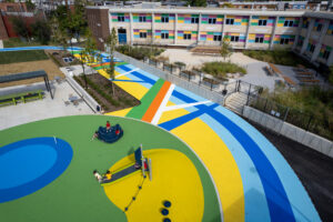 Cecil Community Park: birds-eye view of kids playing on circular playground area featuring green, yellow, and blue poured-in-place rubber paving surrounded by diagonal bands and curving paths of pavement art in blue, turquoise, yellow, white, orange, and green.