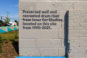 2700 Art Space ministage concrete wall with painted lettering stating "Preserved wall and recreated drum riser from inner Ear Studios, located on this site from 1990-2021."